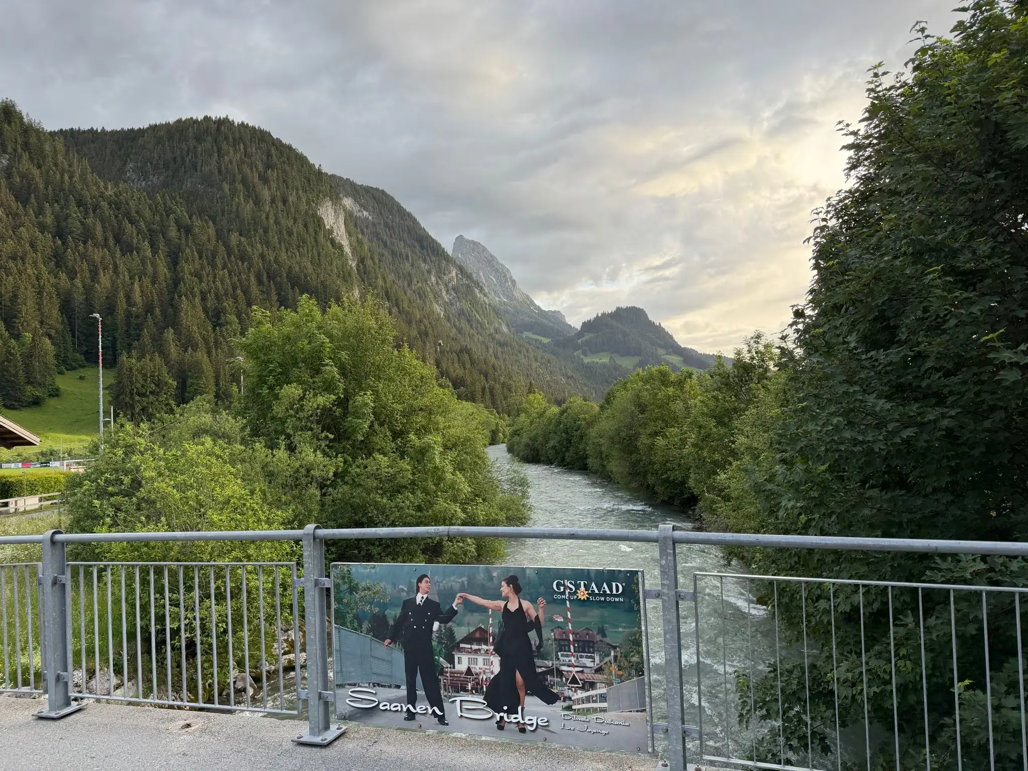 The Saanen Bridge sign referencing the DDLJ mustard-field scene, with the Gstaad valley river and mountains behind